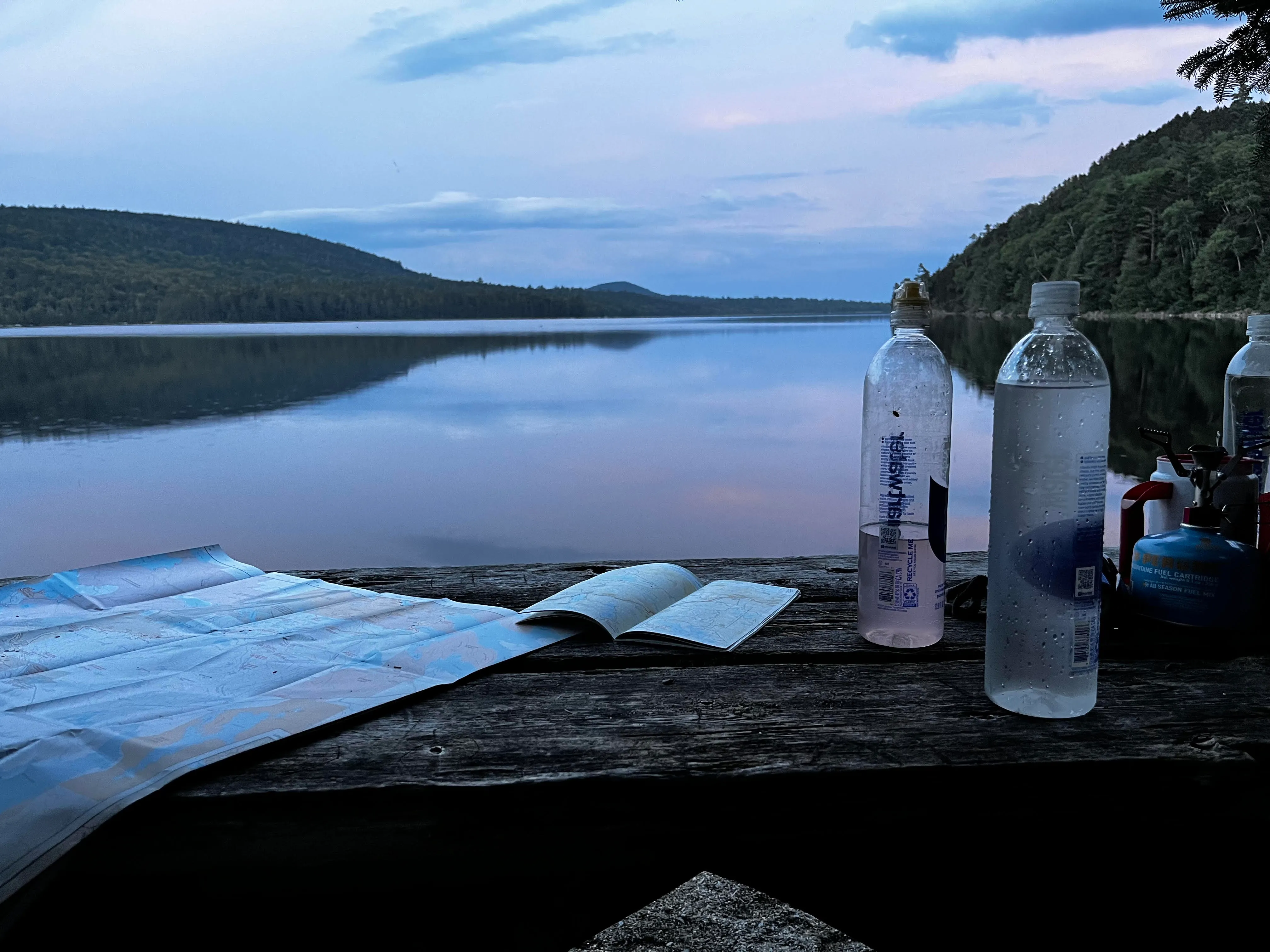 Maps and water bottles spread across a picnic table by a lake at sunset